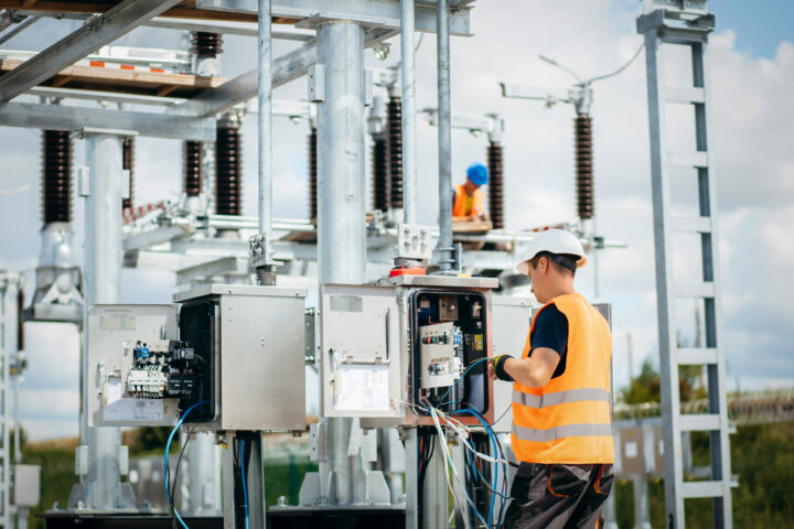 Electrical working on open power box outdoors. Worker is wearing an orange high vis vest and white hard hat.